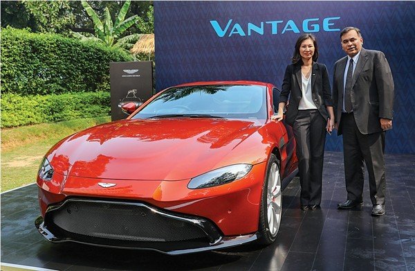 a women and a man standing next to a red color Aston Martin Vantage car 